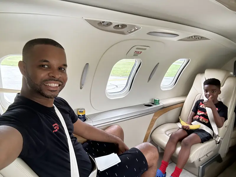 Merrick Lee Jr. smiling in a selfie inside a jet cabin, with Maddox in a cream leather seat behind him