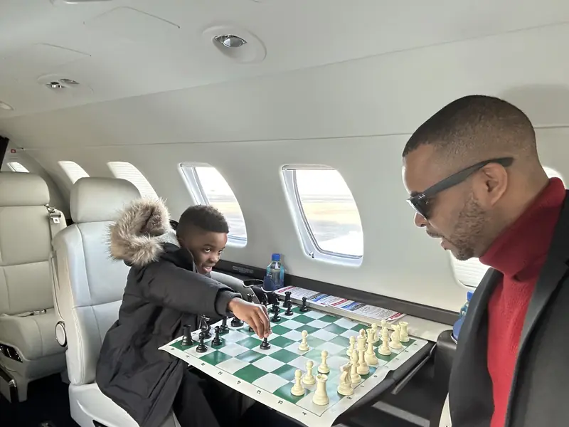 Merrick Lee Jr. and Maddox playing chess at a table inside a private jet cabin