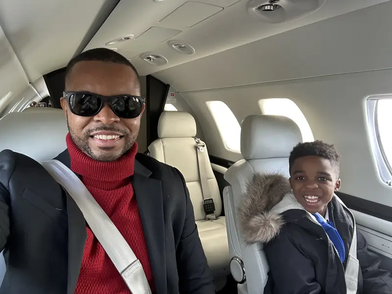 Merrick Lee Jr. smiling in sunglasses and a red turtleneck in a private jet cabin, with Maddox grinning from the seat beside him