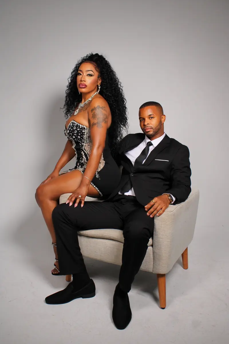 Studio portrait of Merrick Lee in a black tuxedo seated in an armchair, with his partner in a sequined dress beside him